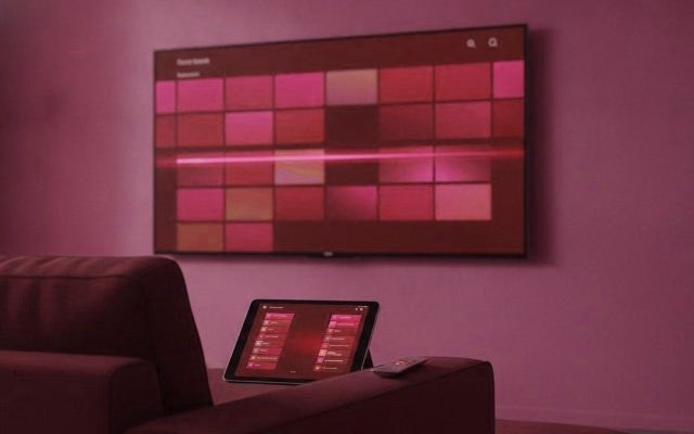 A TV showing a programme guide grid with a tablet and phone showing channel lists on a sofa
