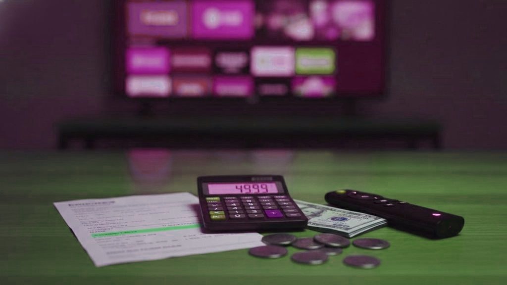 A calculator, dollar bills, coins, a streaming stick and a remote on a table in front of a TV