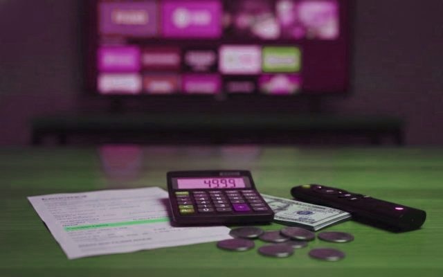 A calculator, dollar bills, coins, a streaming stick and a remote on a table in front of a TV